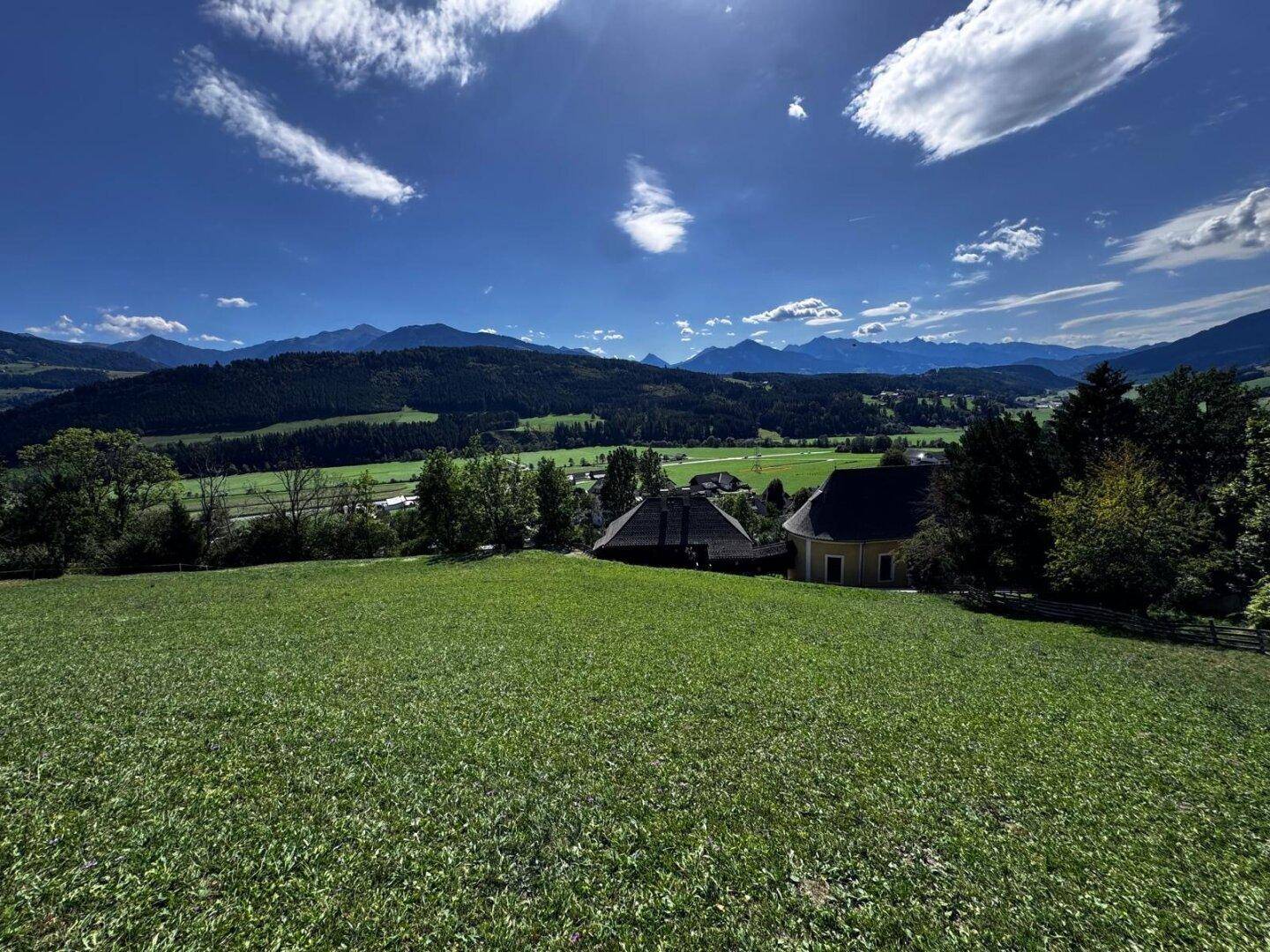 Weitläufige grüne Wiese mit Blick auf ein charmantes Dorf und eine beeindruckende Bergkette unter blauem Himmel.