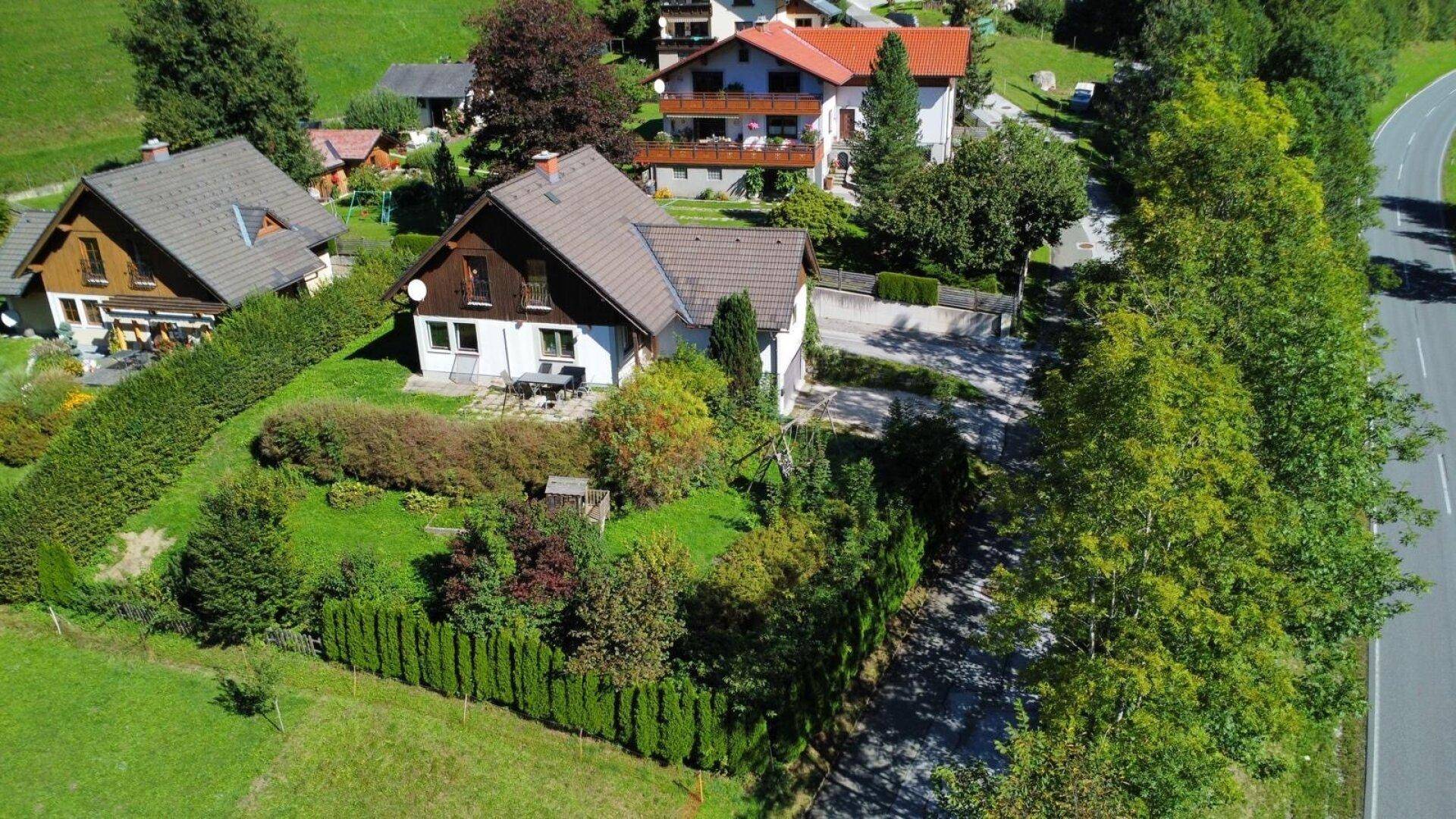 Luftaufnahme eines Hauses mit Garten und angrenzender Straße in einer grünen Berglandschaft.