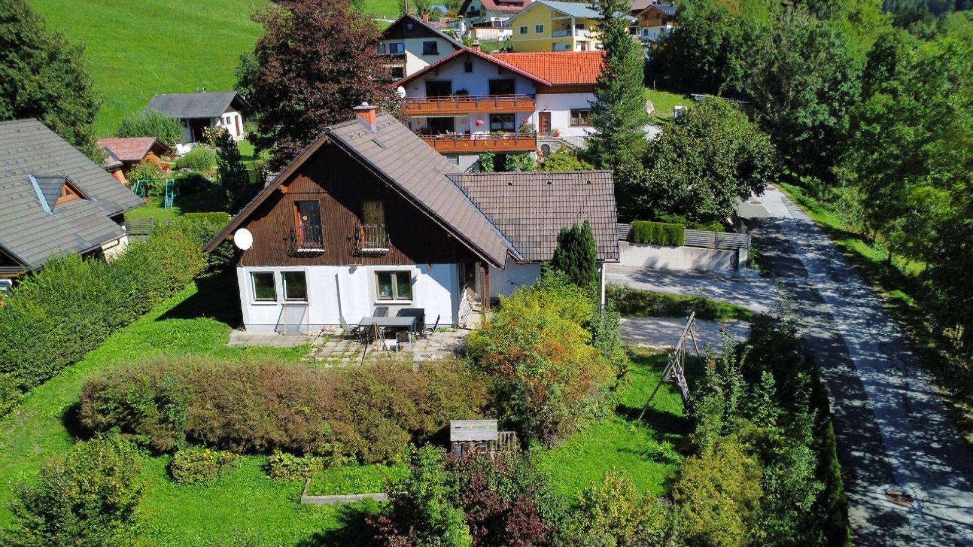 Haus mit kleiner Terrasse und Gartenmöbeln, umgeben von gepflegtem Grün und Bäumen.