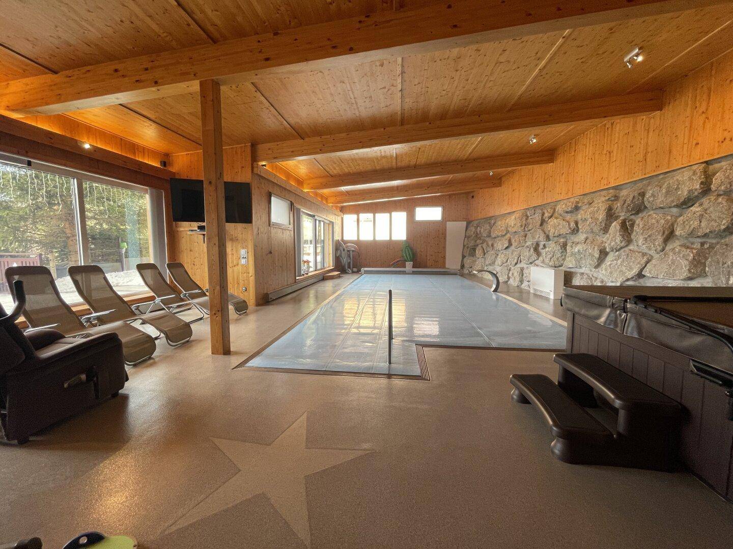 Geräumiger Innenbereich mit überdachtem Pool, Whirlpool und Liegestühlen, umgeben von Holz und Stein.