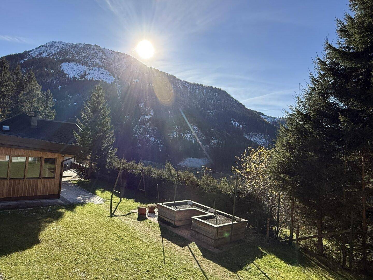 Sonnige Gartenanlage mit Rasen, Bäumen, Hochbeeten und Spielplatz vor verschneiten Bergen.