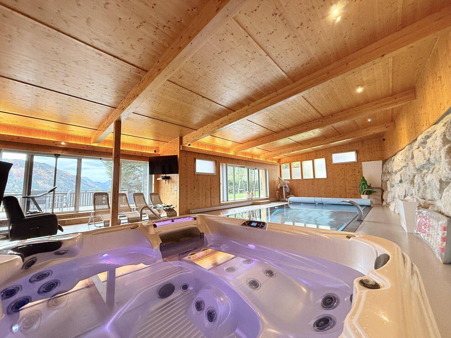Beleuchteter Whirlpool und überdachter Pool in einem rustikalen Wellnessbereich mit Bergblick.
