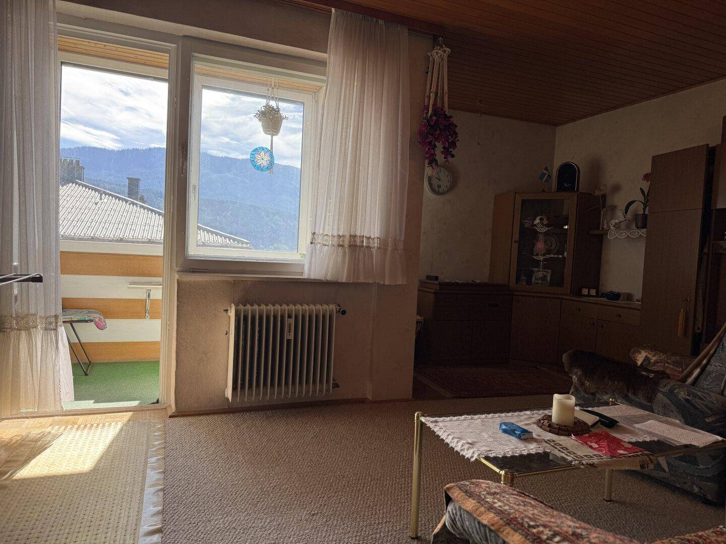 Gemütliches Wohnzimmer mit Teppichboden, Holzdecke und Zugang zu einem kleinen Balkon mit Bergblick.