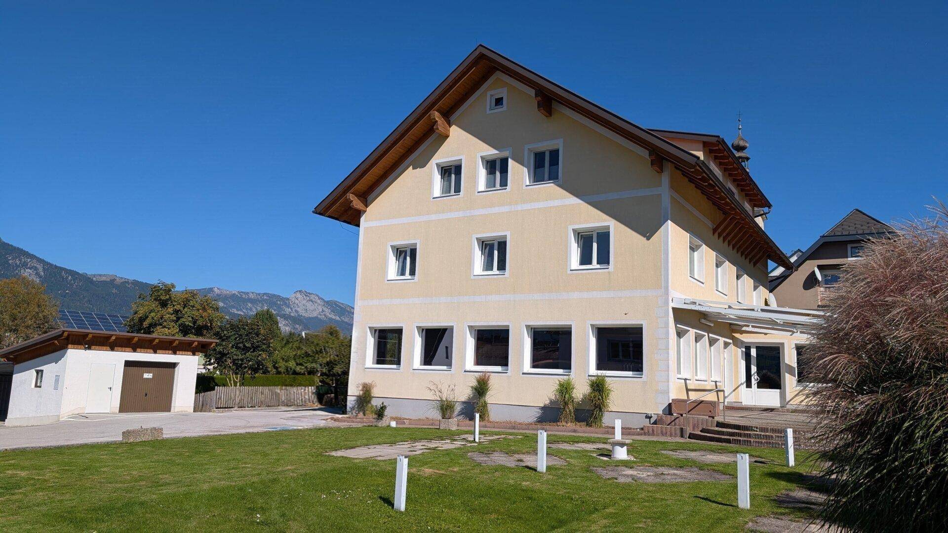 Gelbes Wohngebäude mit Satteldach, umgeben von Rasen und einem Nebengebäude mit Solaranlage.