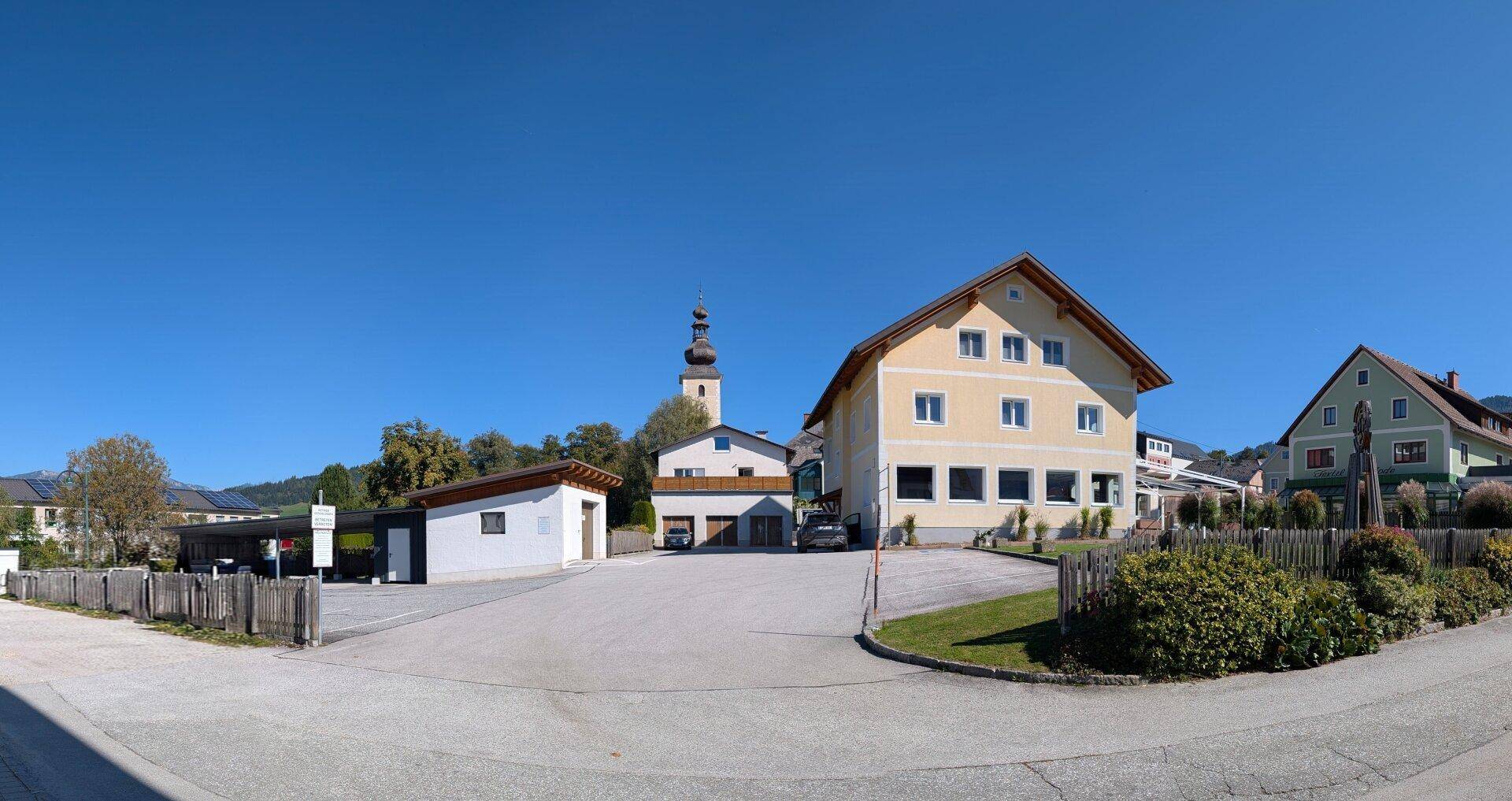 Ansicht des Gebäudes mit Parkplätzen und einem Kirchturm im Hintergrund unter blauem Himmel.