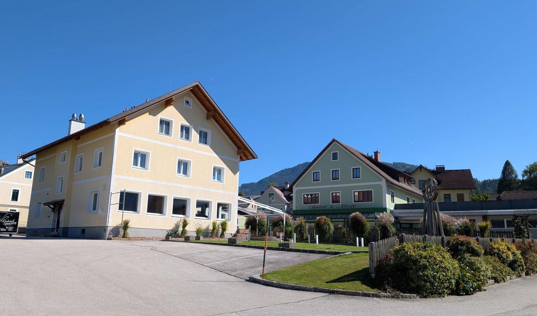 Gelbes Wohngebäude mit Parkplatz und einem weiteren Gebäude im Hintergrund, umgeben von Bergen.