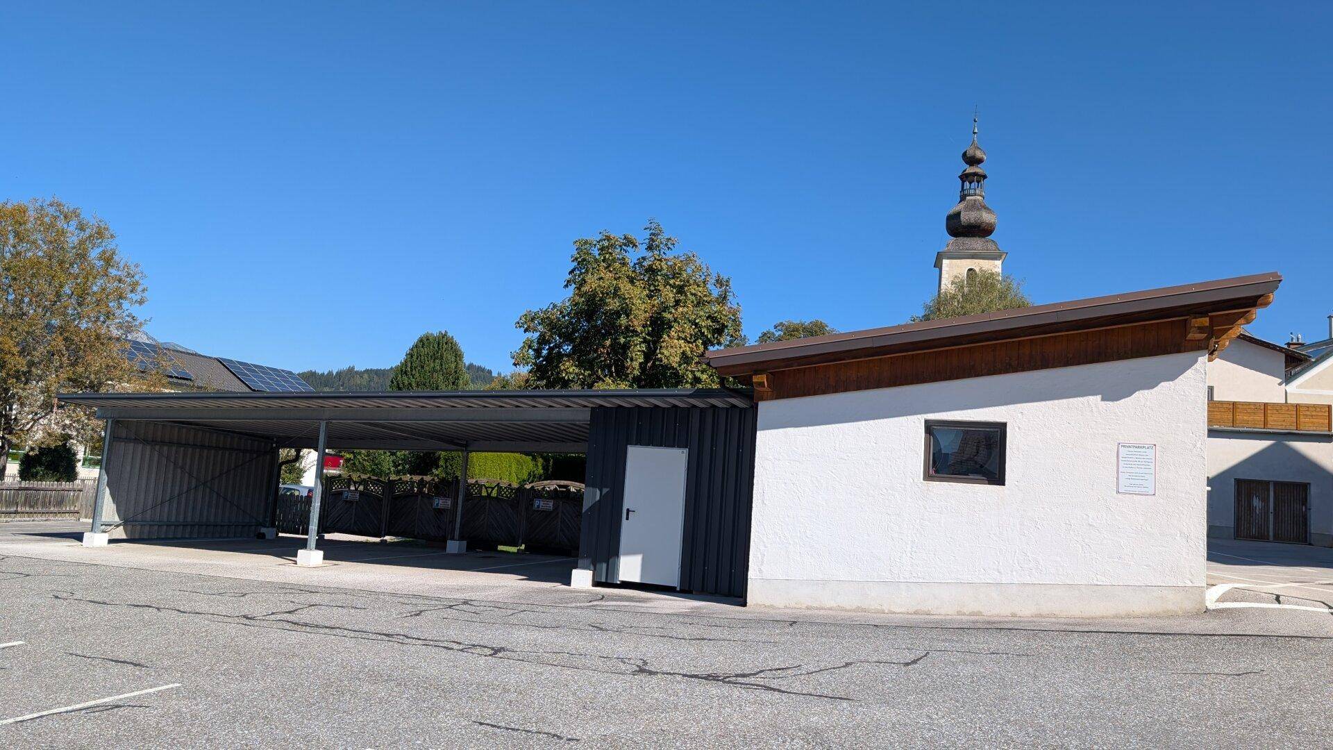 Moderner Carport und ein kleines Nebengebäude mit Kirchturm und Bergen im Hintergrund.