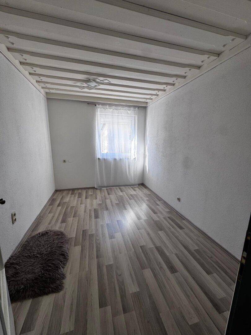 Leeres Zimmer mit Holzboden, weiß gestrichenen Deckenbalken und einem Fenster.