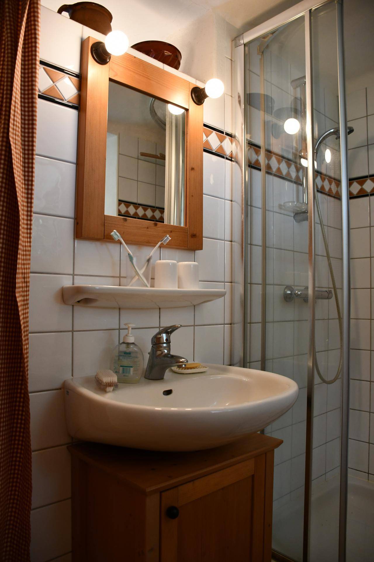 Kleines Badezimmer mit Waschbecken, Holzspiegel und Duschkabine, gefliest in Weiß und Braun.
