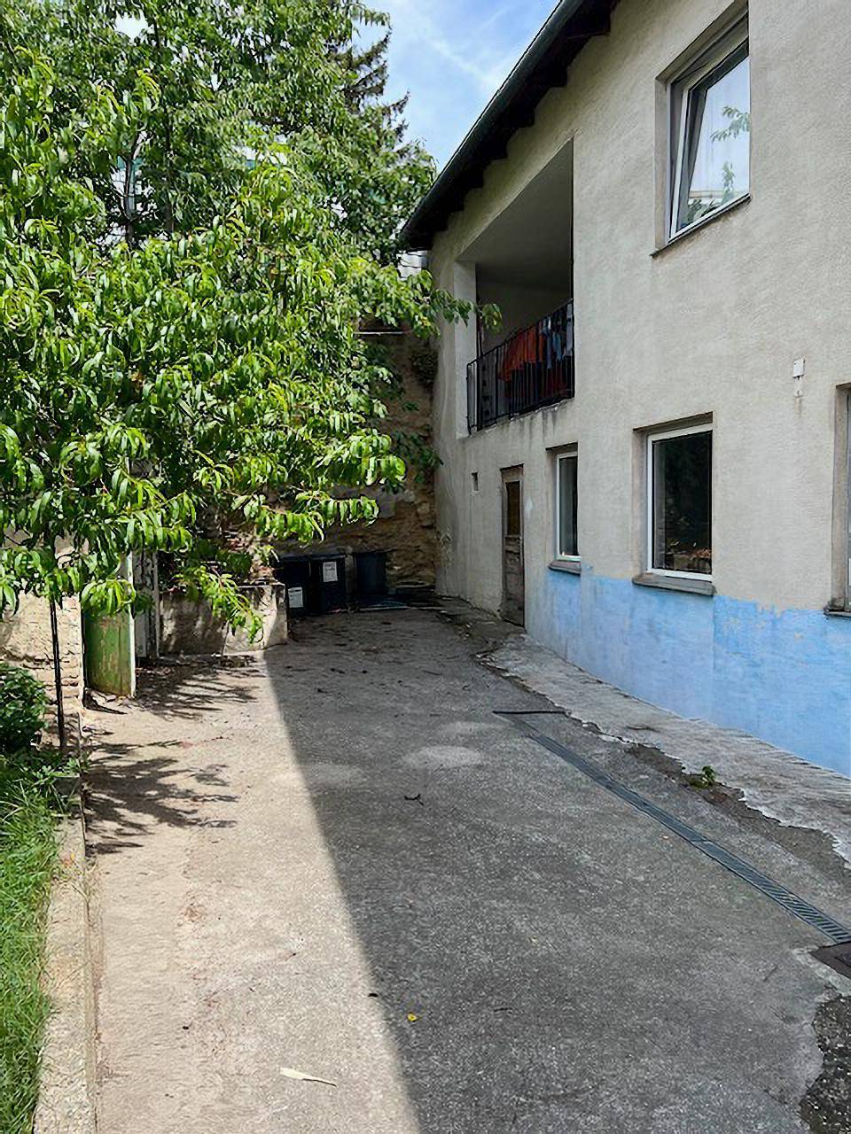 Seitenansicht eines Gebäudes mit schmalem Weg, üppigem Grün und einem kleinen Balkon.