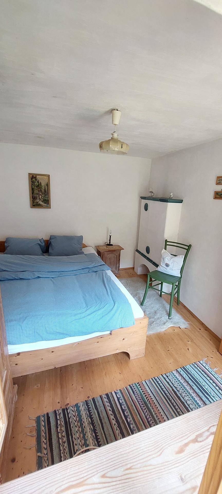 Helles Schlafzimmer mit Holzbett, blauer Bettwäsche, traditionellem Kachelofen und einem farbenfrohen Teppich.