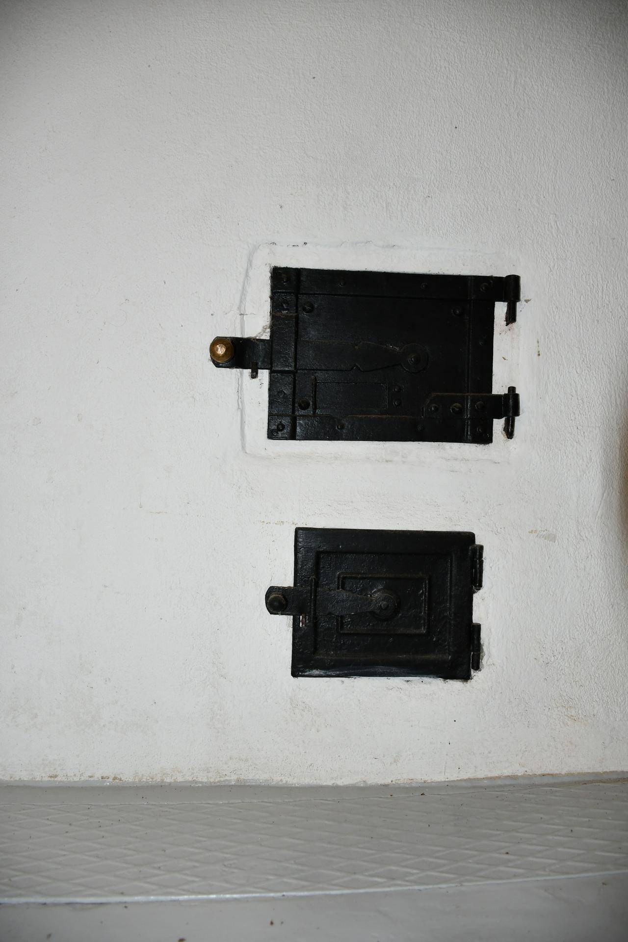 Nahaufnahme von zwei kleinen, schwarzen Metalltüren in einer weißen Wand, die zu einem Ofen gehören könnten.