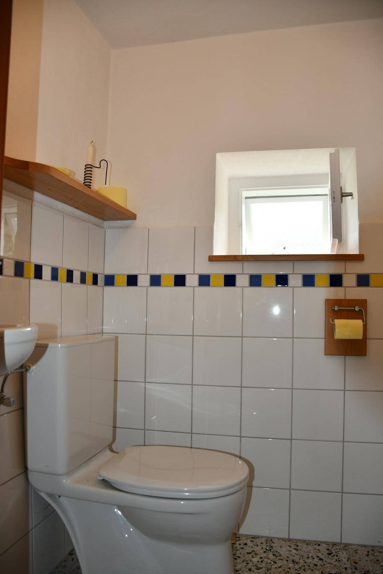 Kleines WC mit weißen Fliesen, farbiger Bordüre und einem Fenster für Tageslicht.