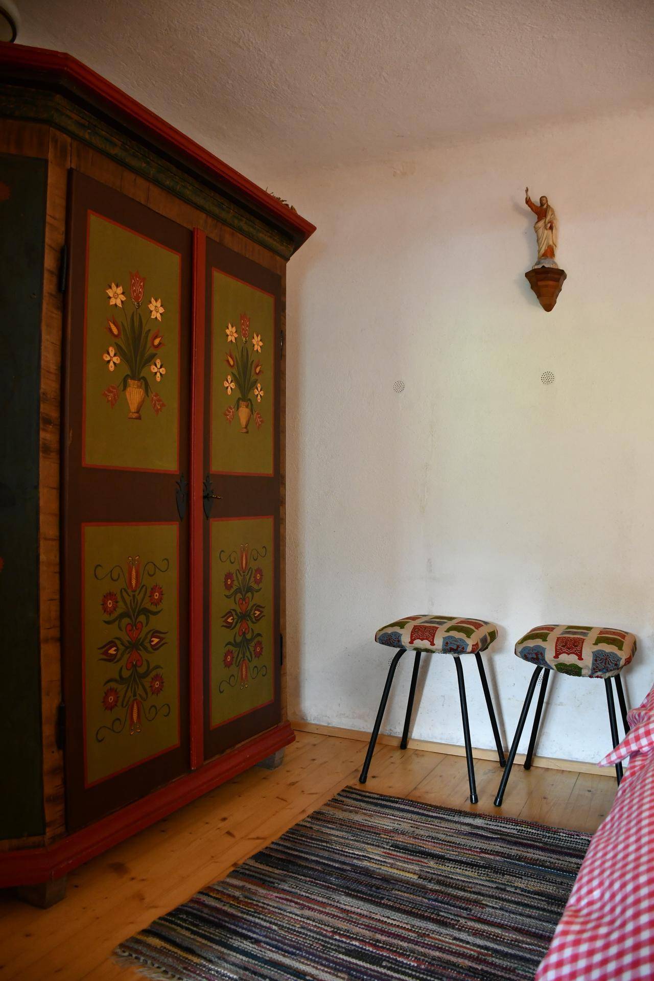 Schlafzimmer mit einem kunstvoll bemalten Kleiderschrank, Holzboden und zwei kleinen Hockern.