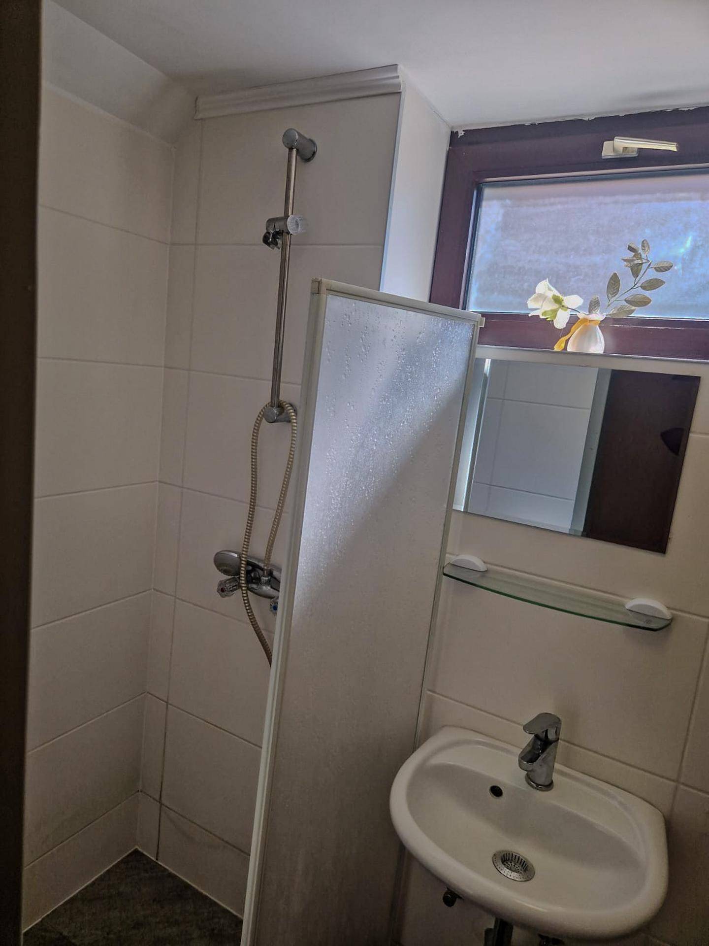 Kleines Badezimmer mit Dusche, Einzelwaschbecken und Fenster, ausgestattet mit weißen Fliesen.