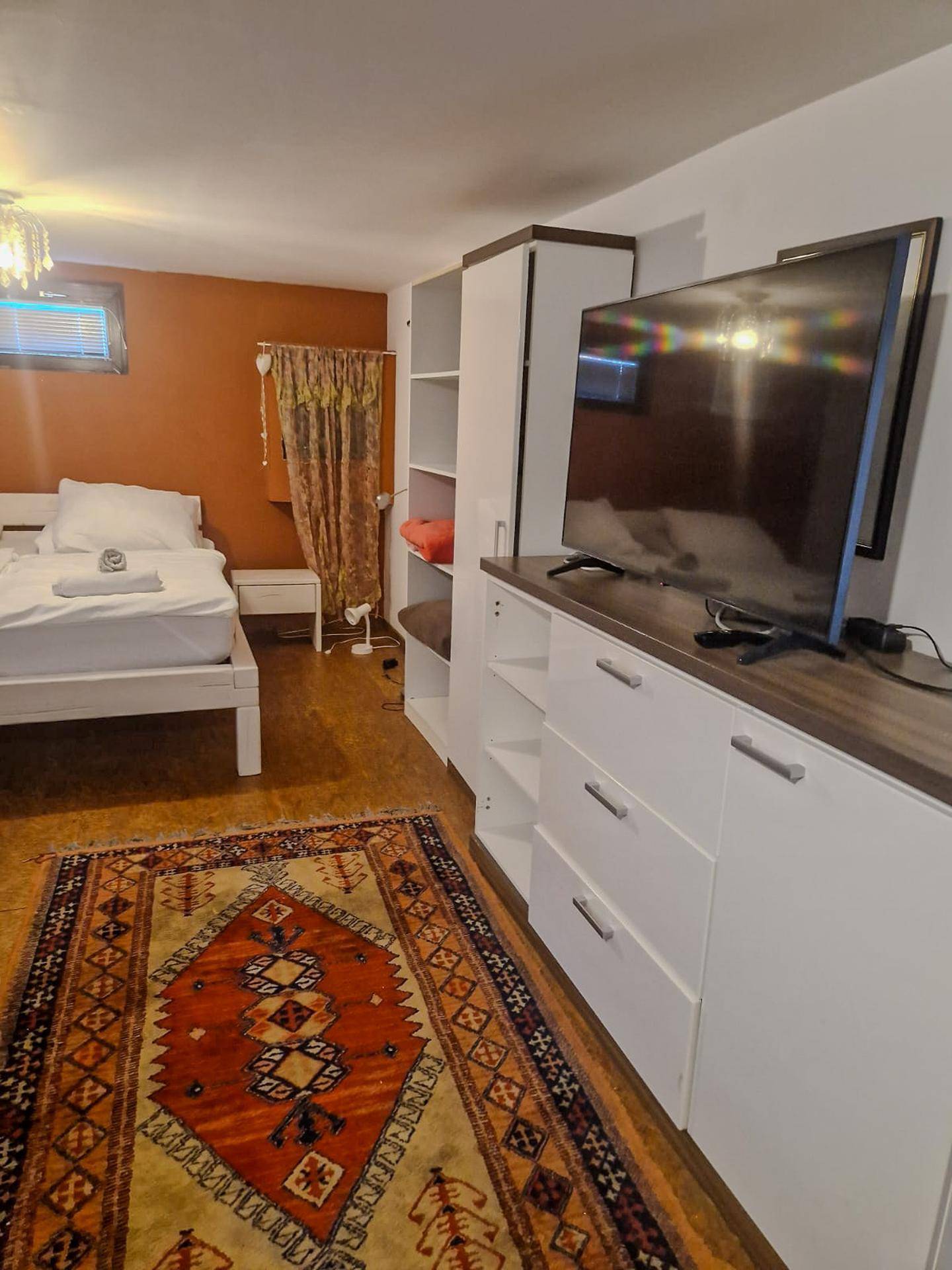 Gemütliches Schlafzimmer mit Doppelbett, Stauraum und Fernseher, akzentuiert durch eine orangefarbene Wand.