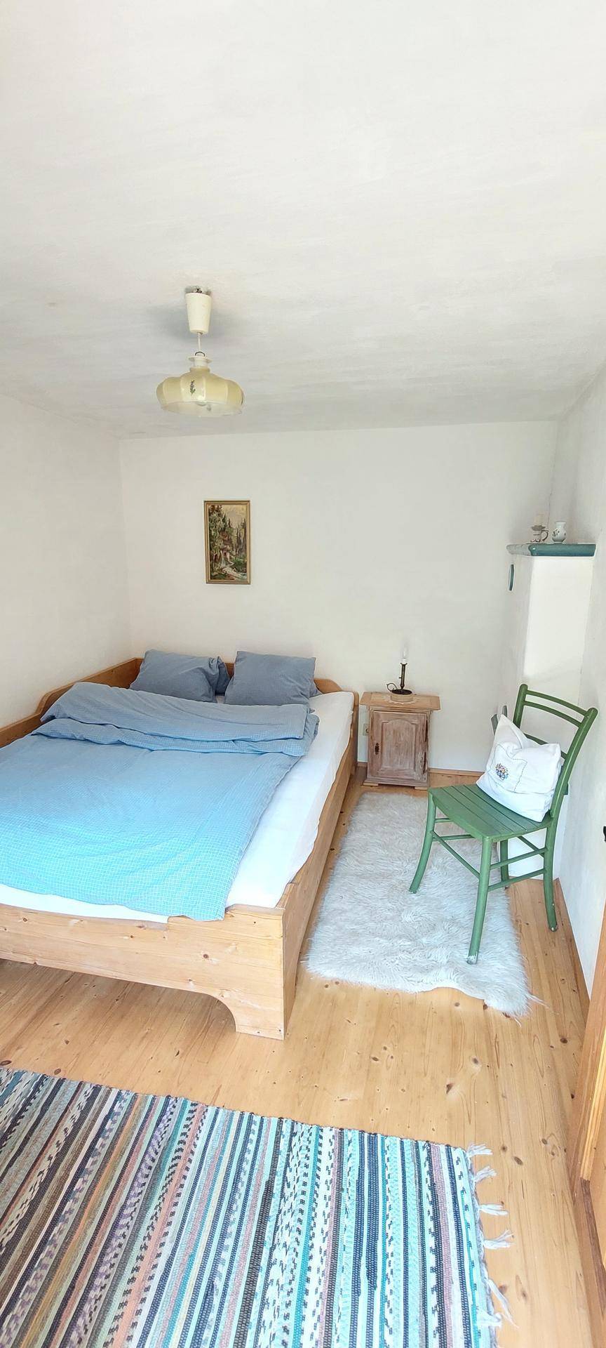 Helles Schlafzimmer mit Holzbett, blauer Bettwäsche, grünem Stuhl und einem farbenfrohen Teppich auf Holzfußboden.