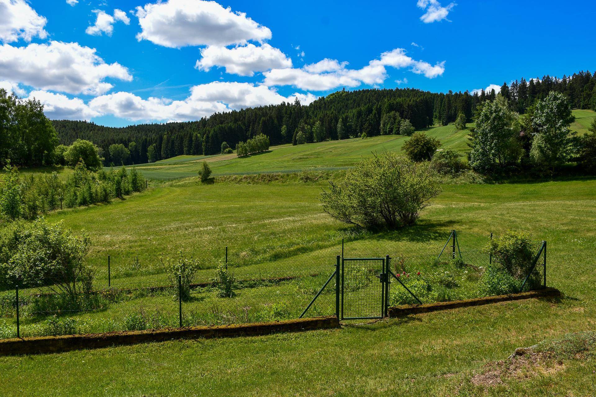 Weitläufiger Gartenbereich mit Zaun, grünen Wiesen und bewaldeten Hügeln im Hintergrund.