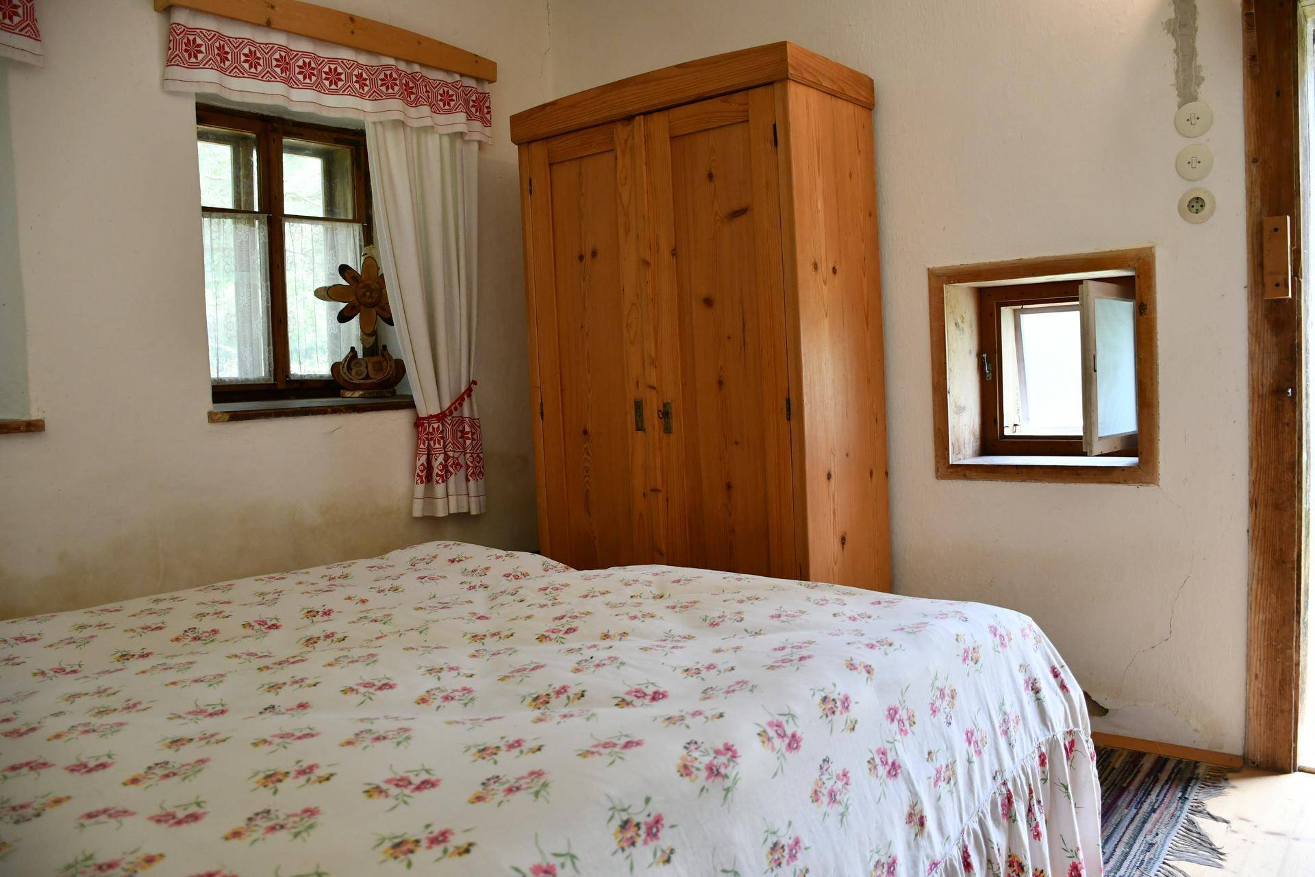 Schlafzimmer mit rustikalem Holzkleiderschrank, floralem Bett und zwei Fenstern mit traditionellen roten Vorhängen.