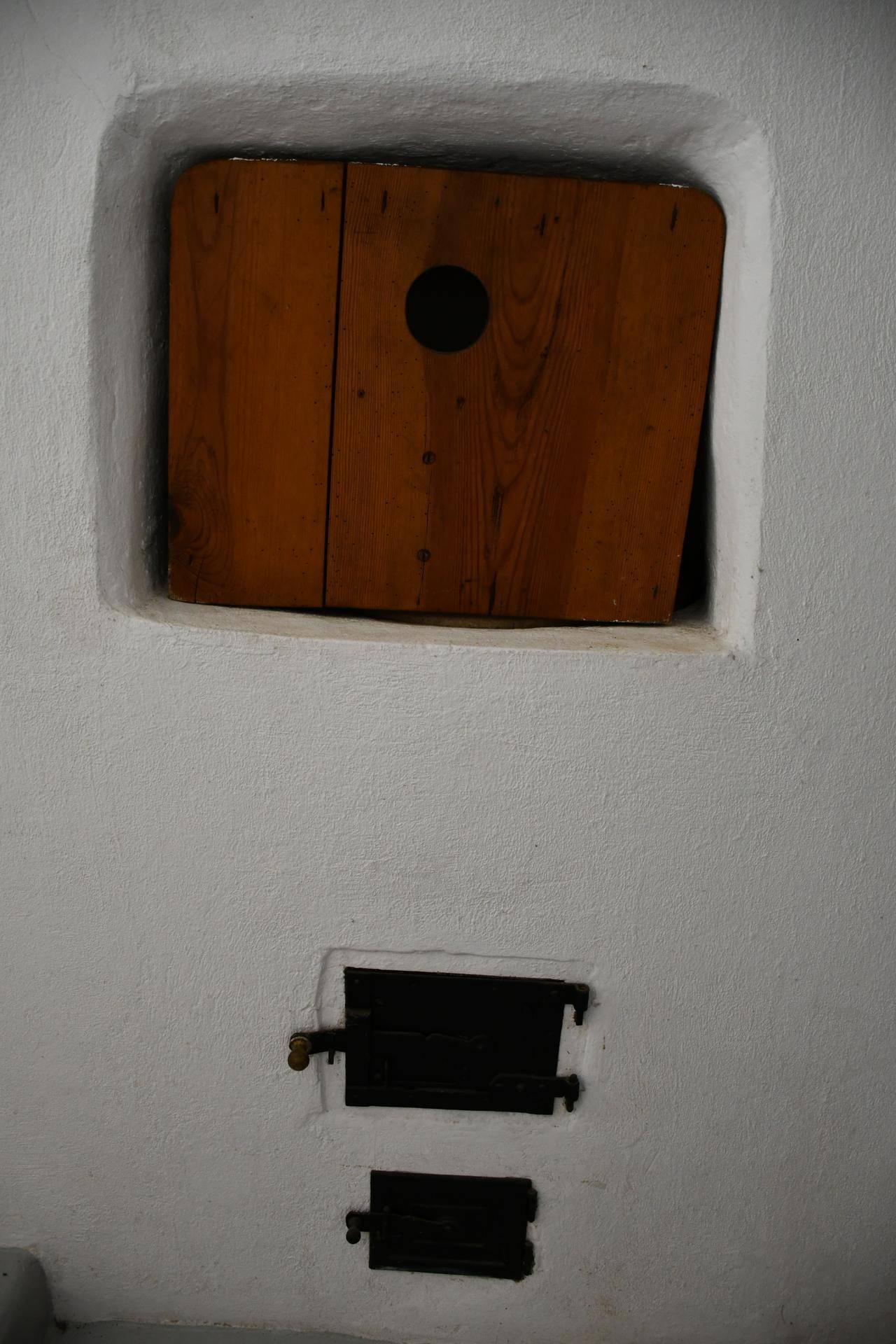 Weiße Wand mit einer Holzluke und zwei kleinen, schwarzen Metalltüren, möglicherweise für einen Ofen.