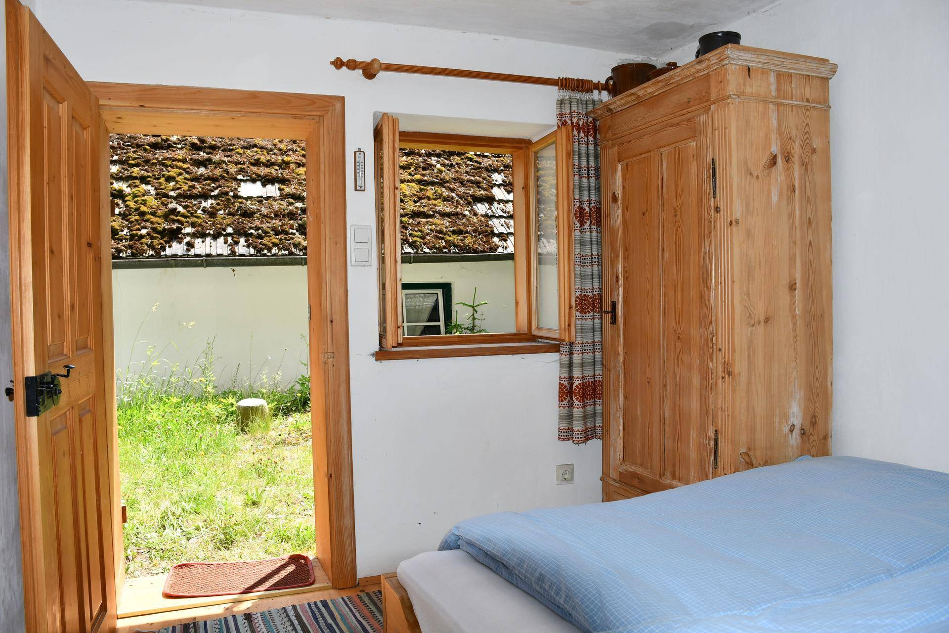 Schlafzimmer mit Holzbett und Schrank, offener Tür und Fenster mit Blick auf einen grünen Außenbereich.