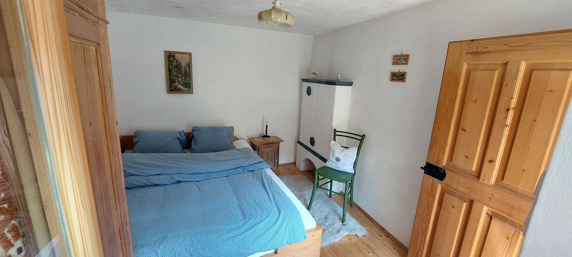 Schlafzimmer mit Holzbett, blauem Bettzeug, Kachelofen und rustikalen Holzmöbeln, die eine warme Atmosphäre schaffen.