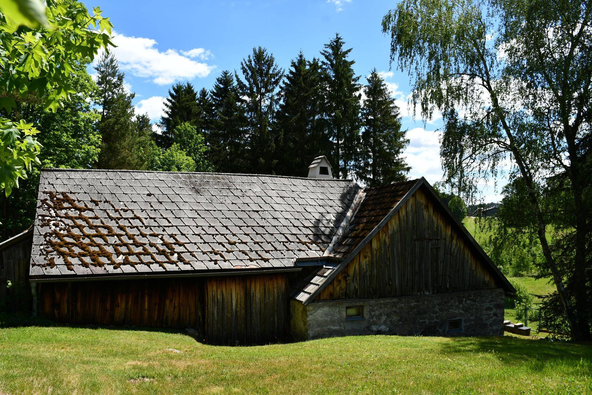 Ländliches Gebäude mit Holzanbau und Steinfundament, umgeben von grünen Wiesen.