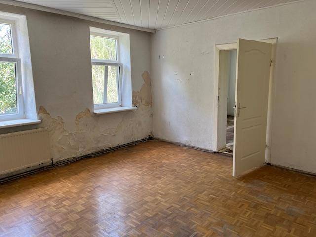 Leerer Raum mit Parkettboden, abblätternder Farbe an den Wänden und zwei Fenstern.