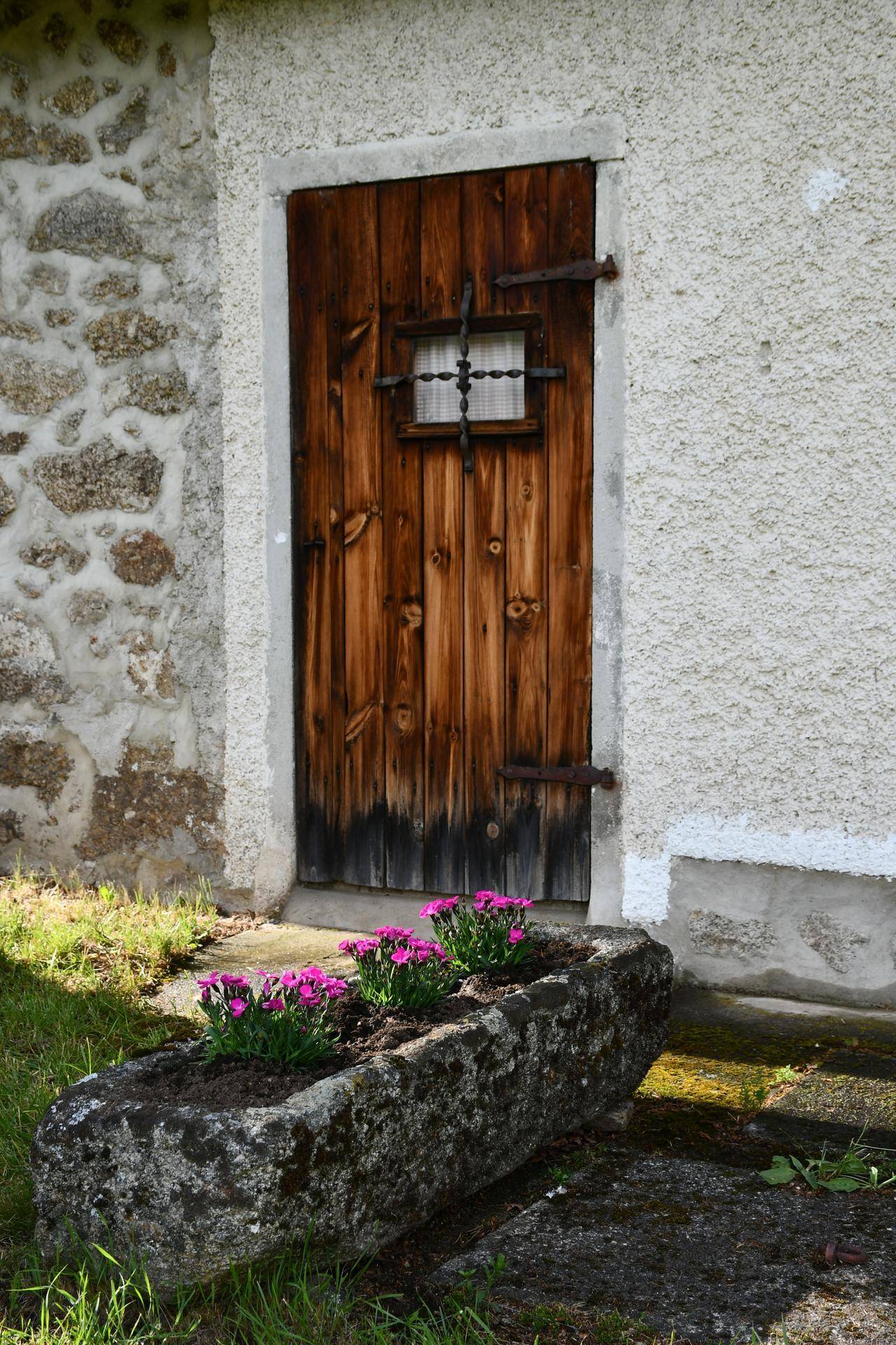 Rustikale Holztür mit kleinem Fenster und schmiedeeisernen Beschlägen, davor ein steinerner Blumenkasten.