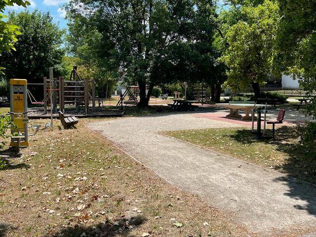 Öffentlicher Spielplatz mit verschiedenen Geräten und Sitzgelegenheiten unter Bäumen.
