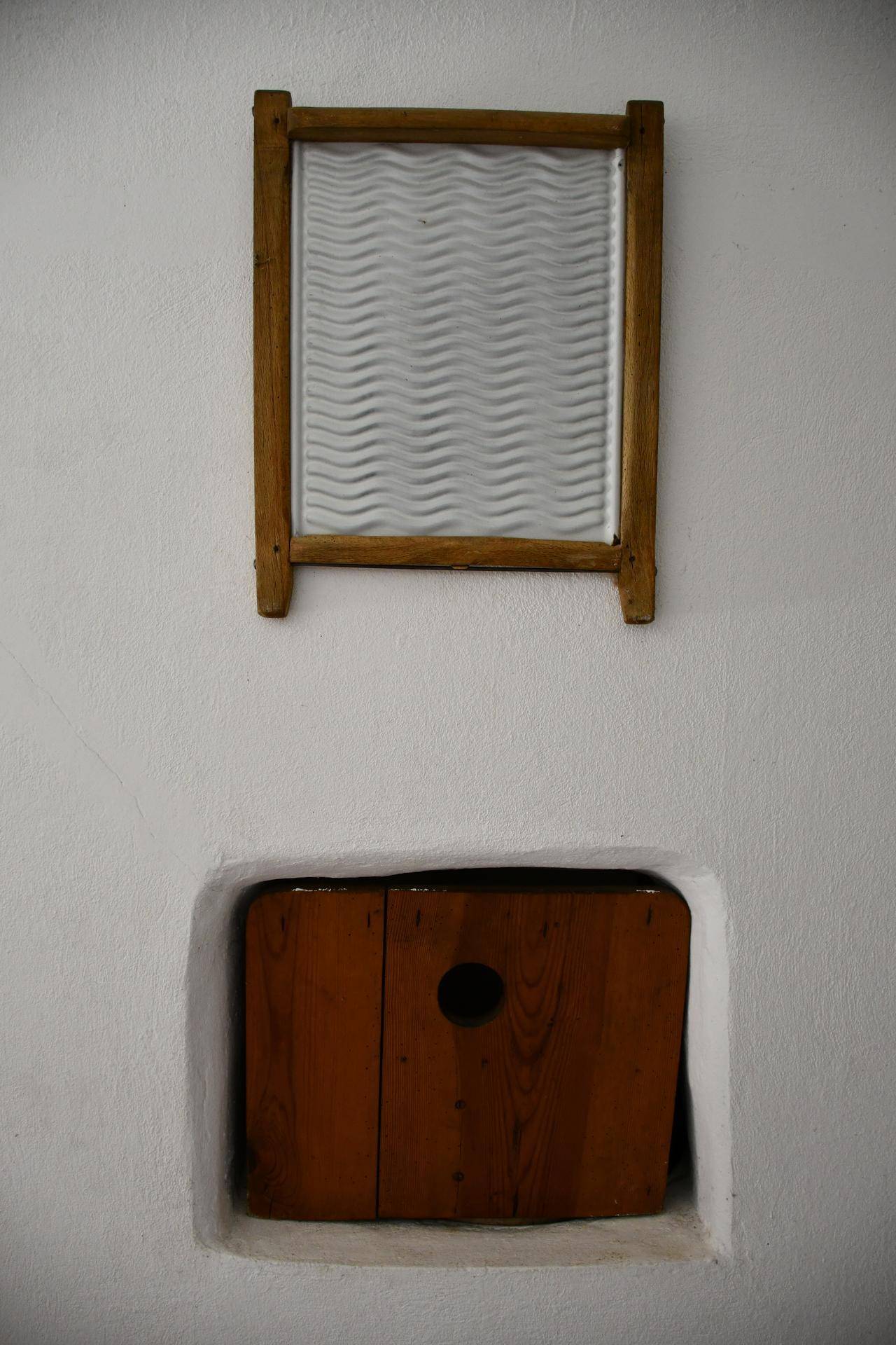 Weiße Wand mit einem alten Waschbrett und einer kleinen Holzluke, die auf traditionelle Nutzung hinweist.