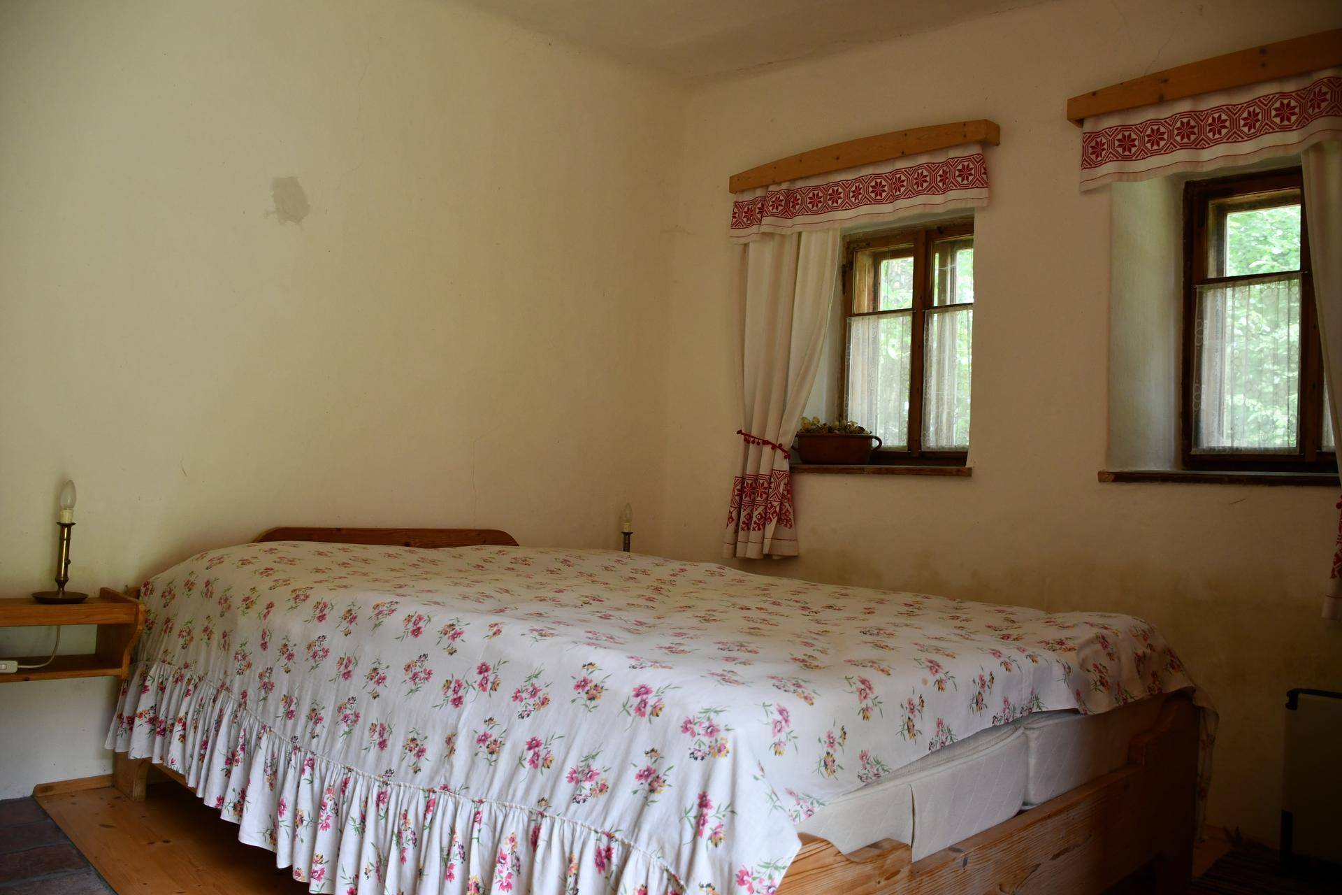 Gemütliches Schlafzimmer mit floralem Bettüberwurf, Holzbett und zwei kleinen Fenstern mit traditionellen Vorhängen.