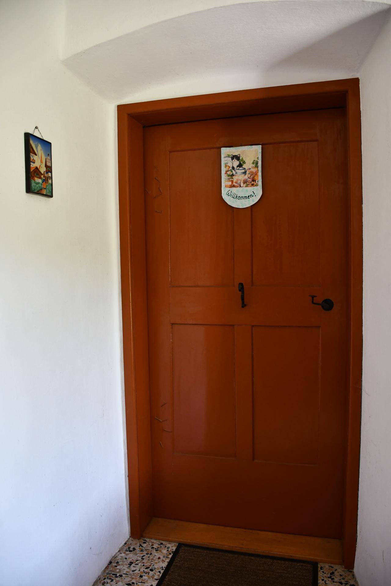 Eine braune Holztür mit einem Willkommensschild und einem kleinen Bild an der weißen Wand.
