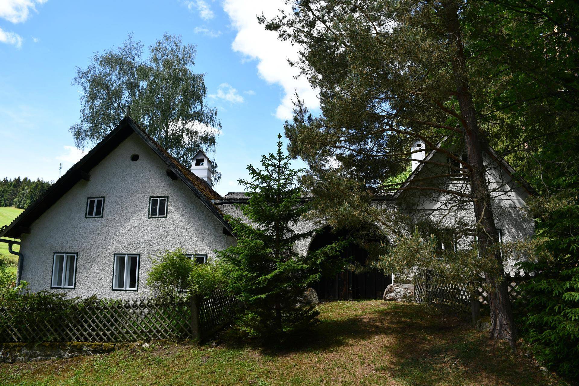 Traditionelles Landhaus mit weißer Fassade und Holzzaun, eingebettet in eine grüne Umgebung.