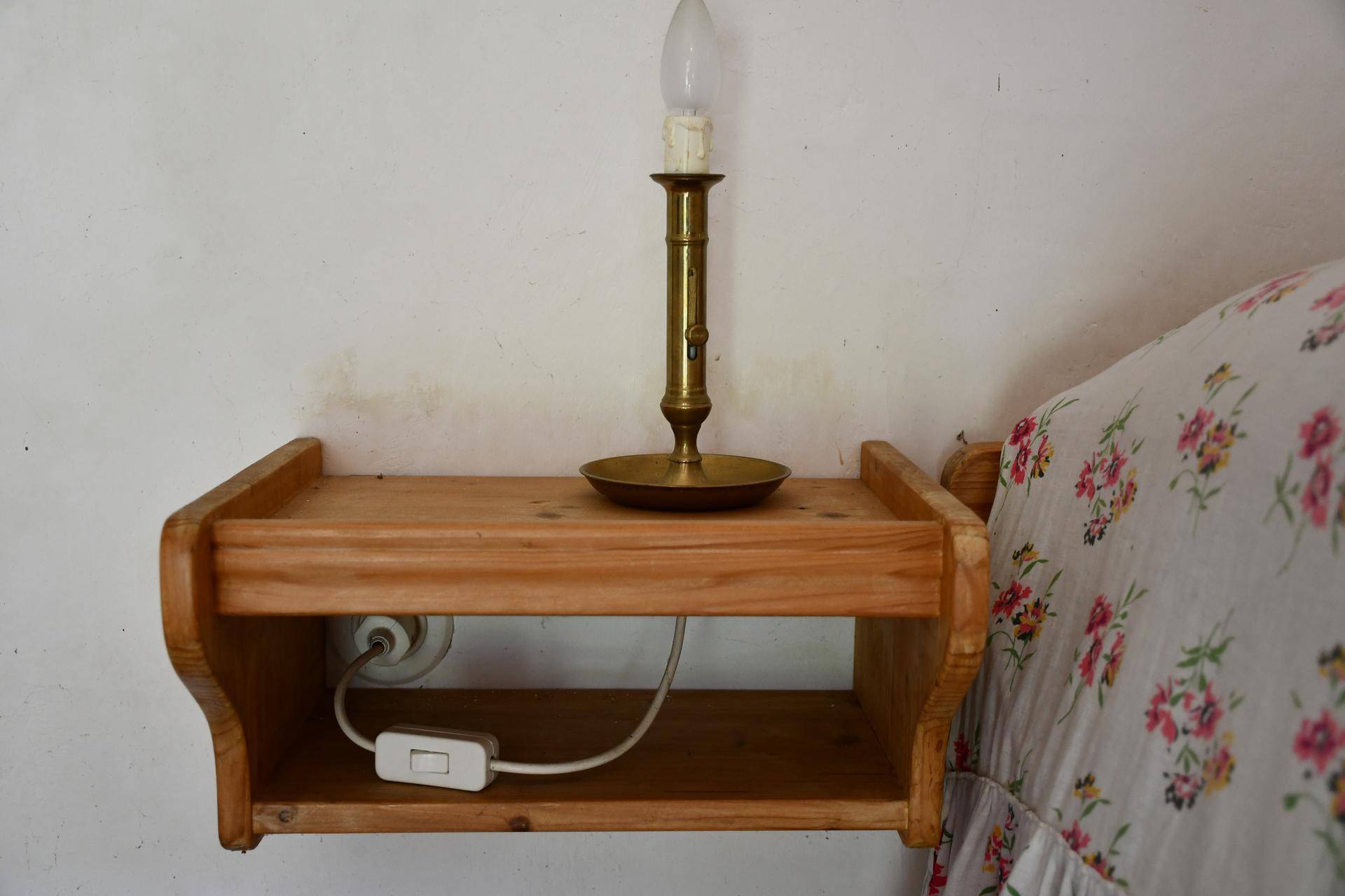 Einfacher hölzerner Nachttisch mit einer klassischen Lampe und floral gemusterter Bettwäsche.