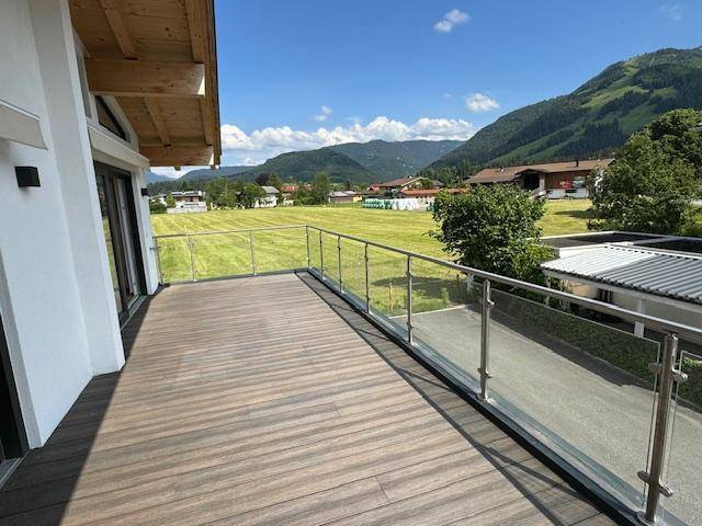 Geräumige Holzterrasse mit Glasgeländer und weitem Blick auf grüne Wiesen und Berge.