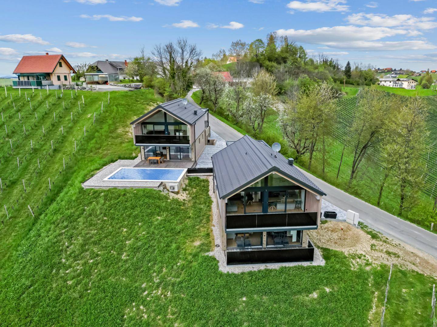 Zwei moderne Häuser mit Pool auf einem grünen Hügel, umgeben von Weinbergen unter blauem Himmel.