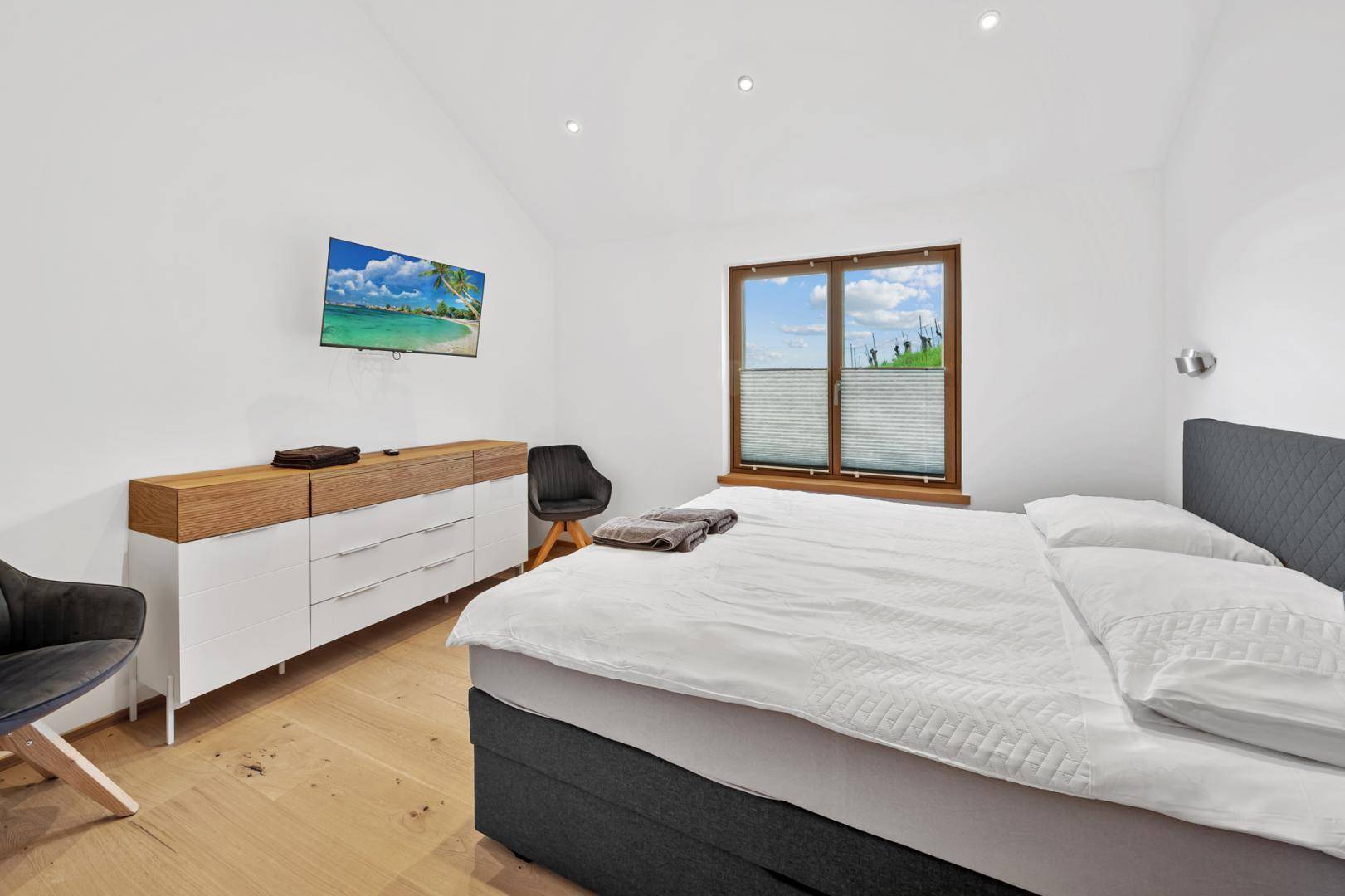 Helles Schlafzimmer mit modernem Bett, Holzkommode und Fenster mit Blick auf die grüne Landschaft.