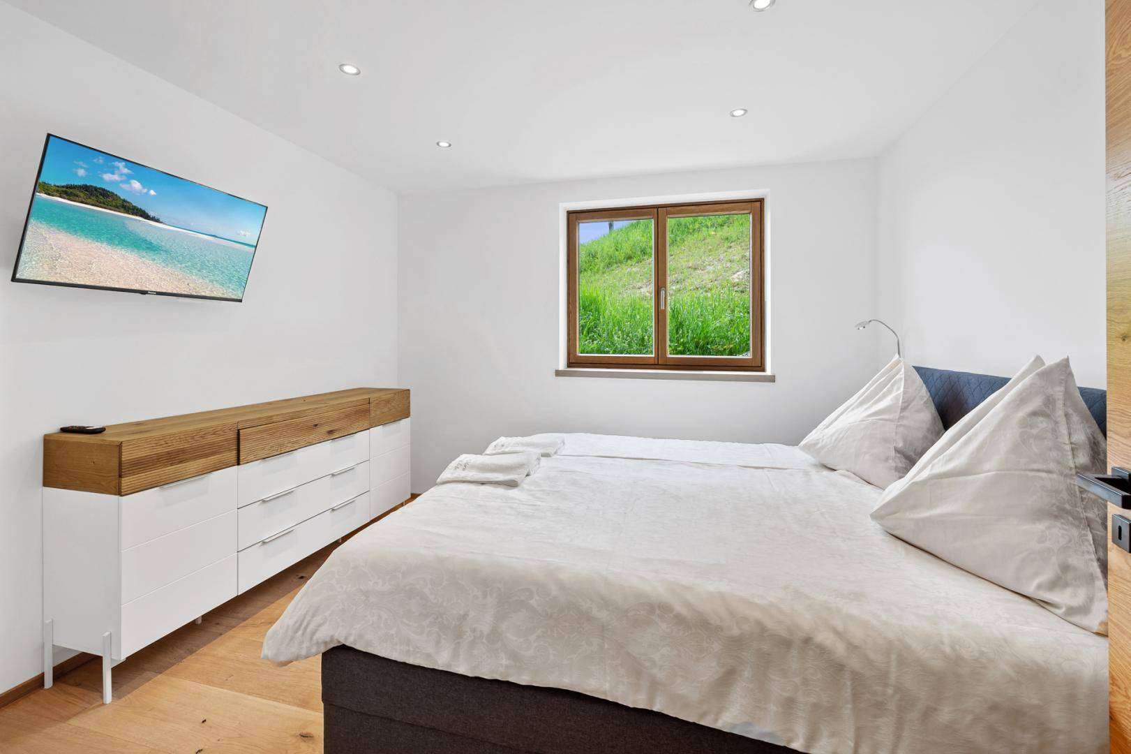 Helles Schlafzimmer mit Doppelbett, Holzboden und Blick ins Grüne durch das Fenster.
