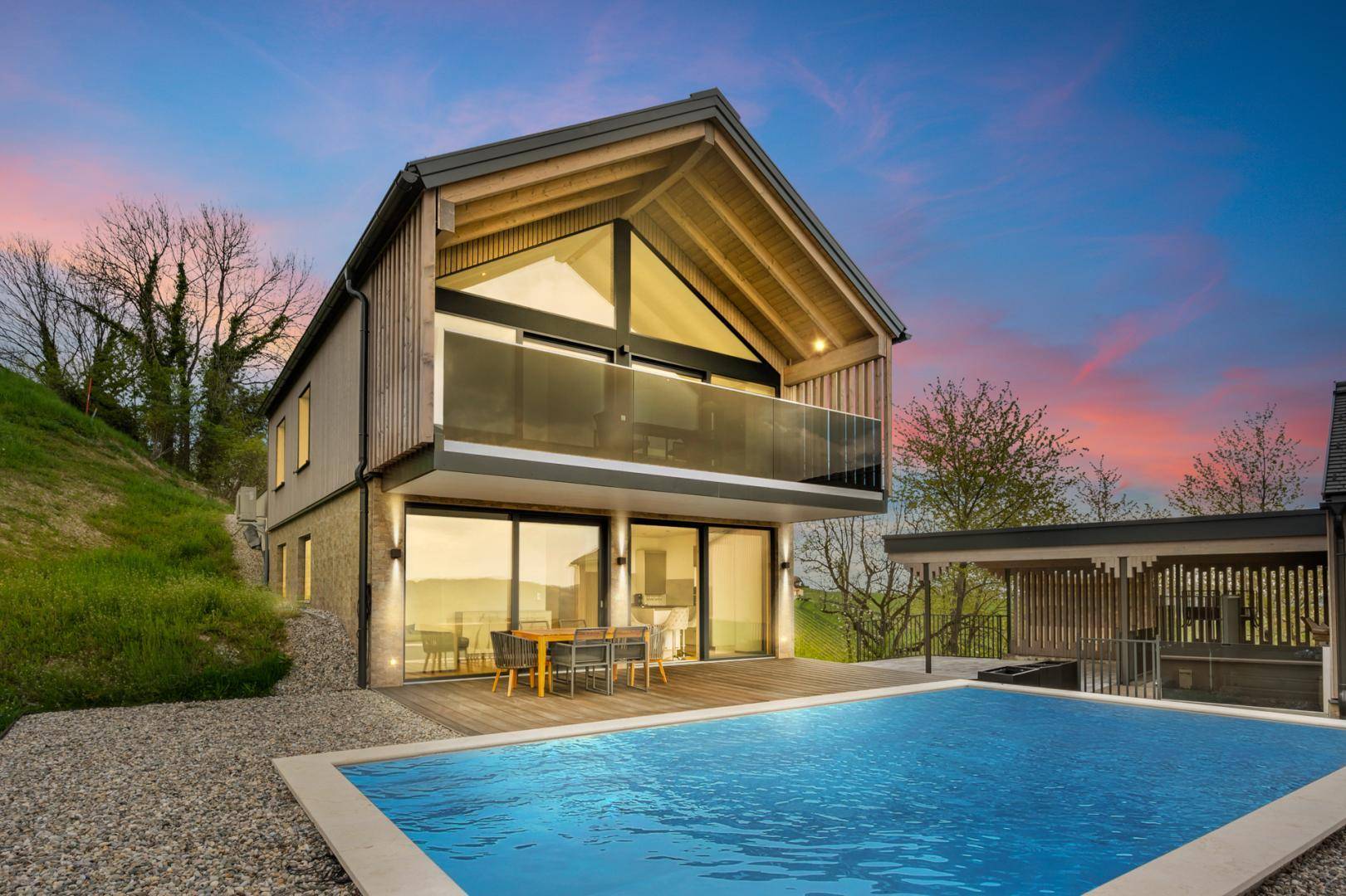 Hinteransicht eines modernen Hauses mit Pool, Holzterrasse und Essbereich im Freien bei Sonnenuntergang.