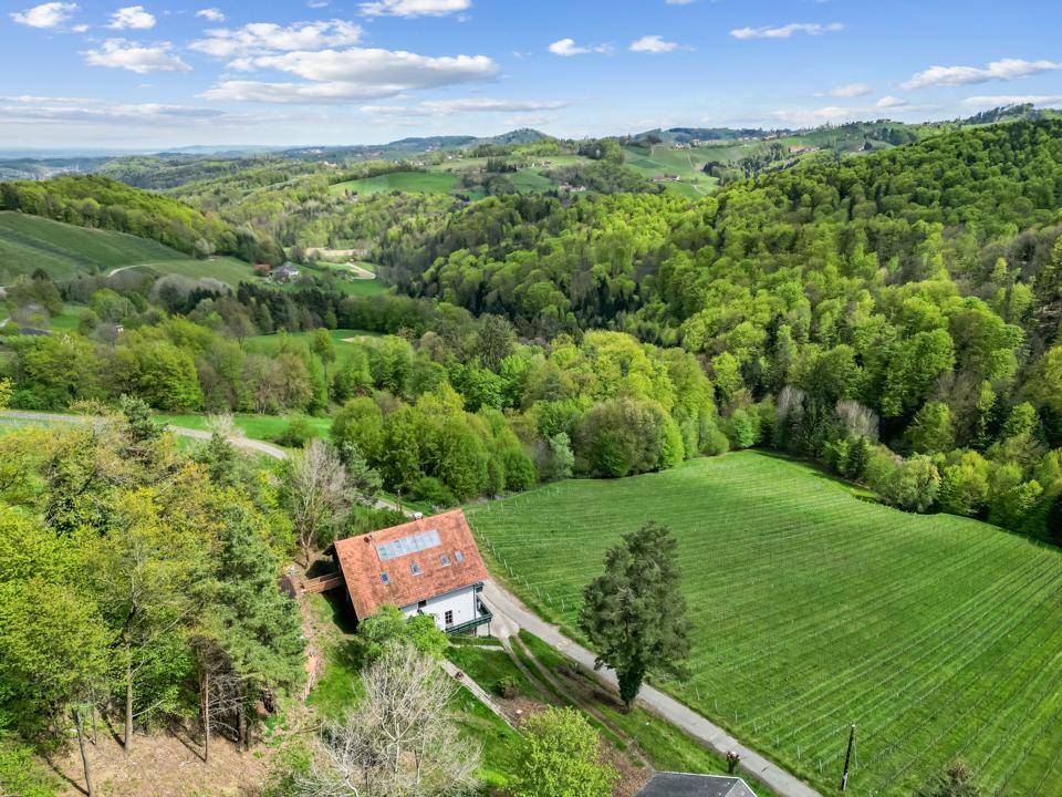 Luftaufnahme eines Hauses eingebettet in eine grüne, hügelige Landschaft mit Wäldern und Feldern.