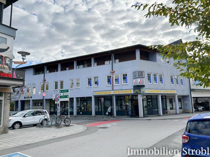 Gebäude an einer Straßenecke mit Geschäften im Erdgeschoss und Wohnungen darüber, umgeben von städtischer Infrastruktur.