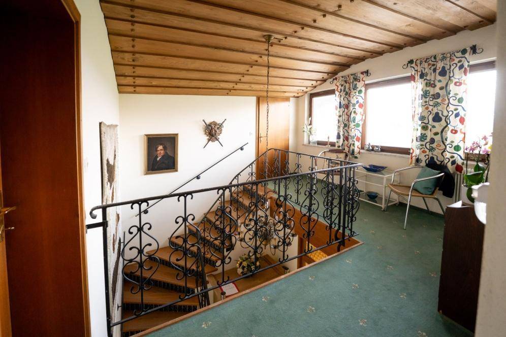 Flur mit traditioneller Holzdecke, schmiedeeisernem Geländer, Teppichboden und Fenstern mit Grünblick.