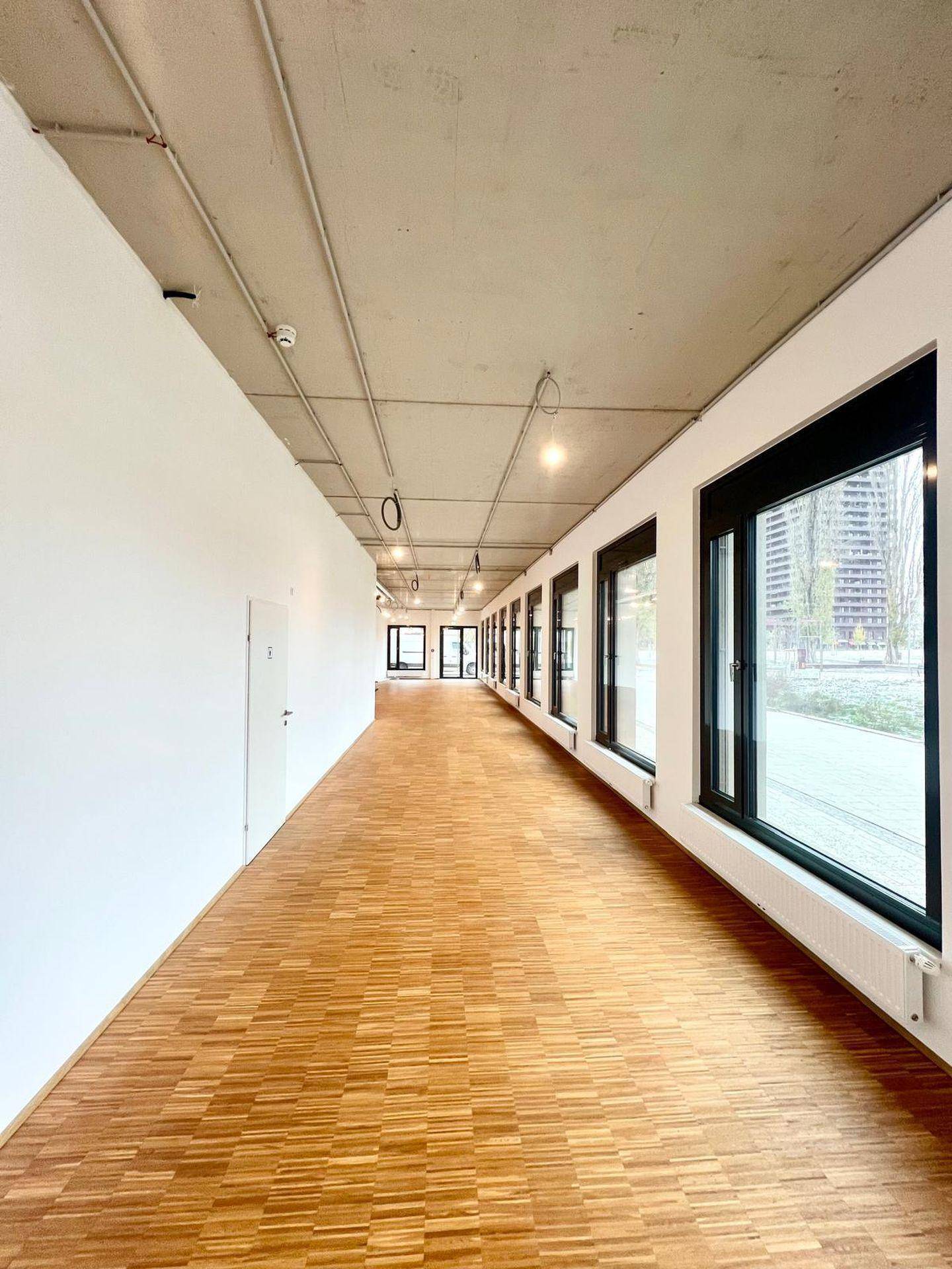 Geräumiger Gewerberaum mit durchgehender Fensterfront, Holzboden und moderner Deckenbeleuchtung in einem Neubau.