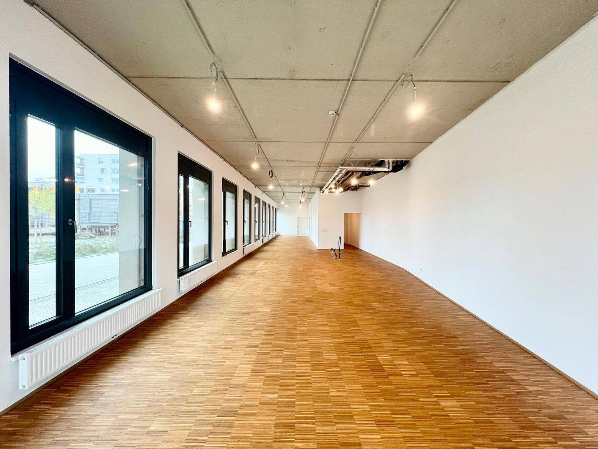 Langer, heller Gewerberaum mit großen Fenstern, Holzboden und Sichtbetondecke, bereit zur individuellen Gestaltung.