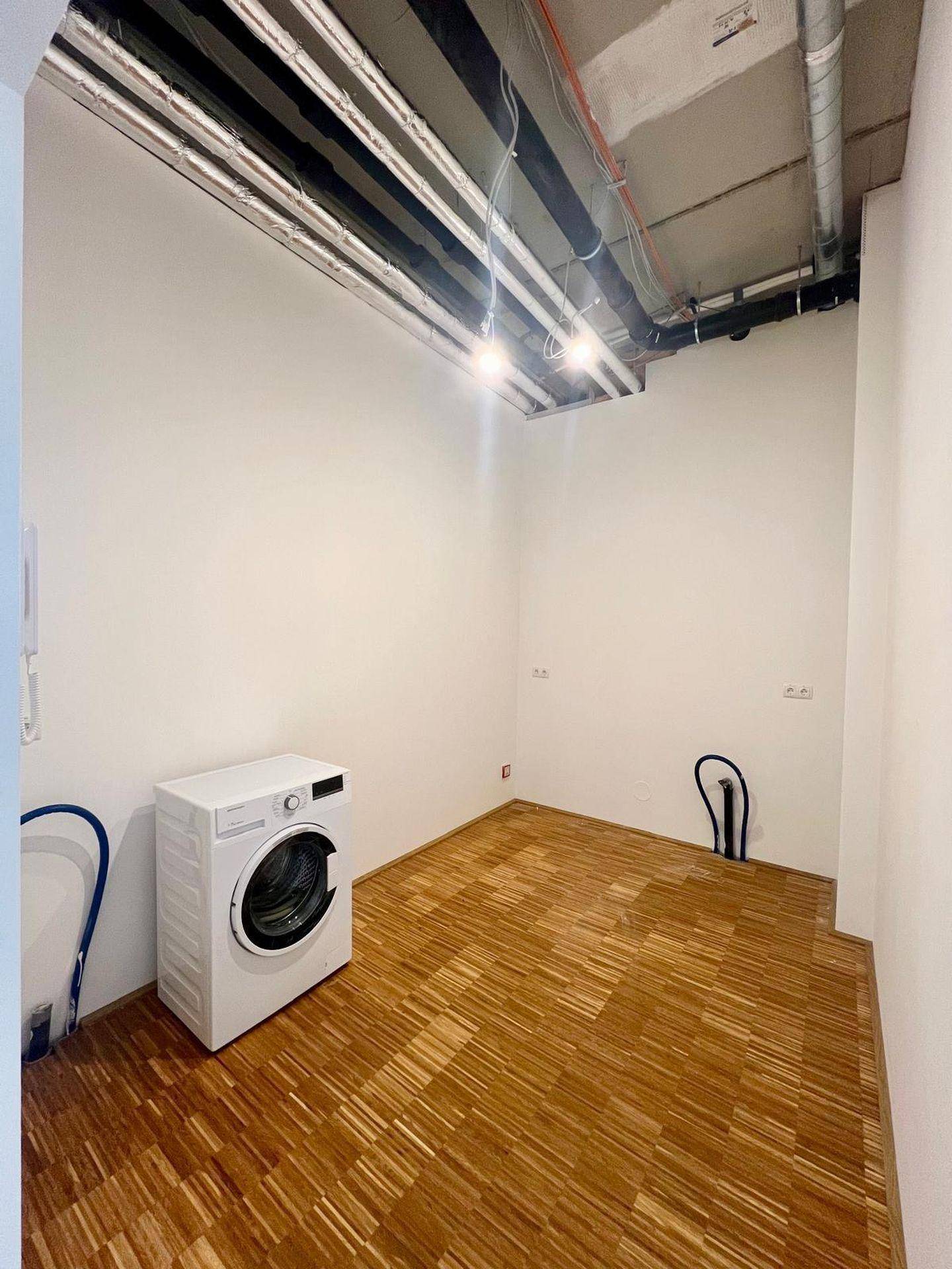 Kleiner Waschraum mit einer Waschmaschine, Holzboden und sichtbaren Leitungen an der Decke.