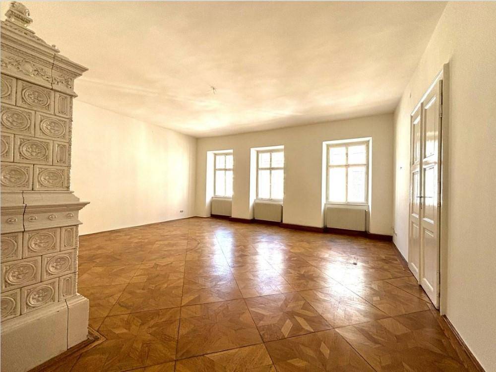 Geräumiges Wohnzimmer mit traditionellem Kachelofen, Fischgrätparkett und hohen Fenstern, das viel natürliches Licht erhält.