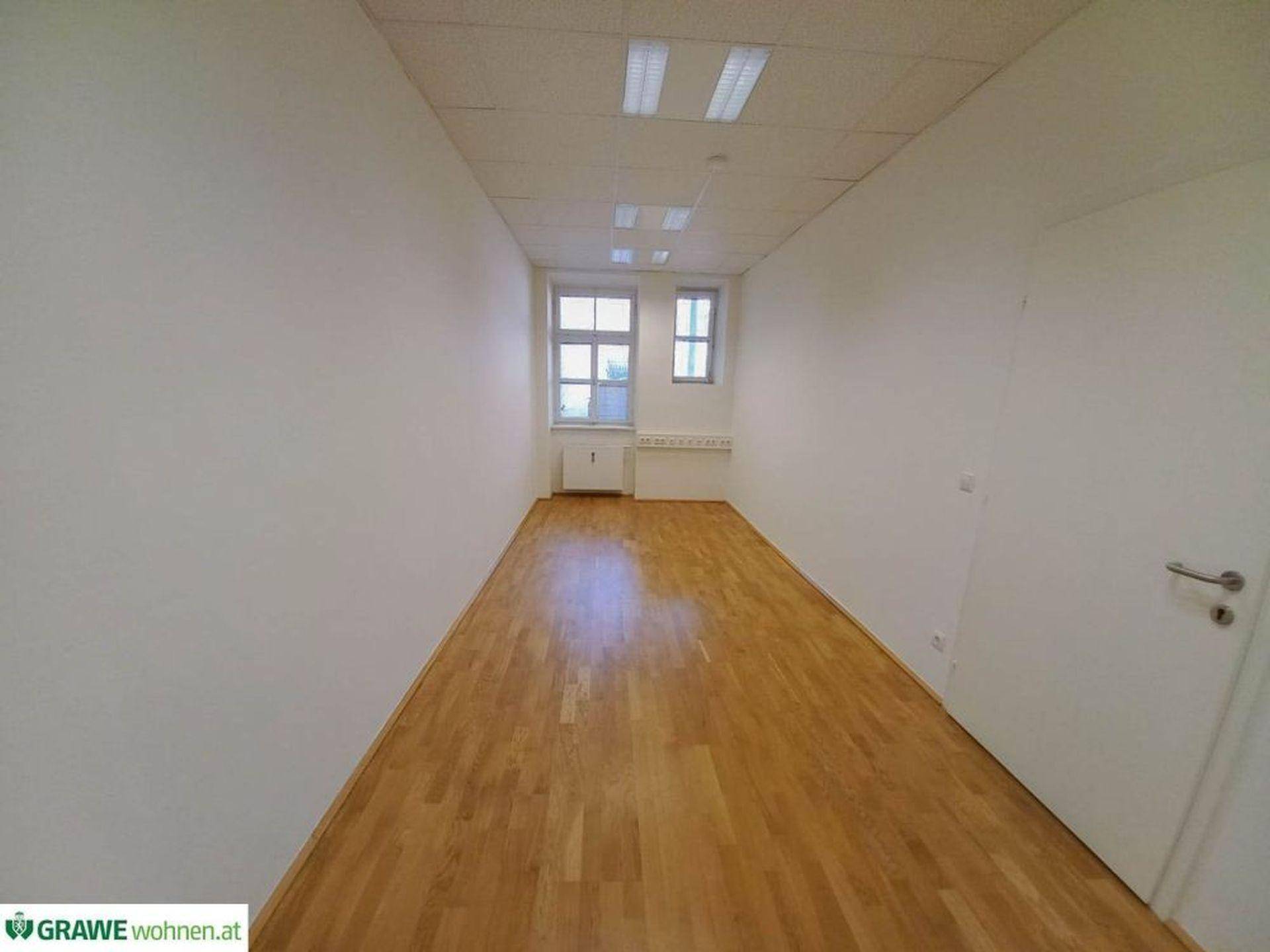 Langes, schmales Büro mit hellem Holzfußboden und Fenstern am Ende des Raumes, ideal für Arbeitsplätze.