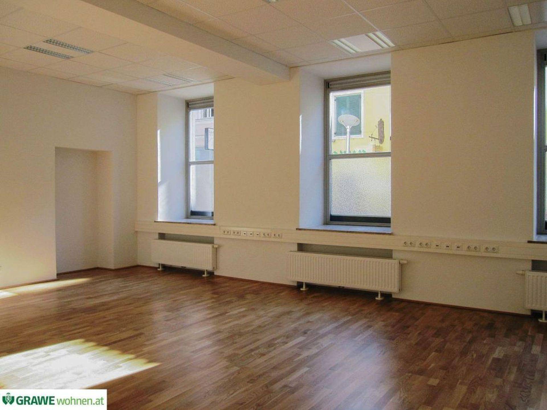 Leeres Büro mit hellem Holzfußboden, weißen Wänden und Heizkörpern unter den großen Fenstern.