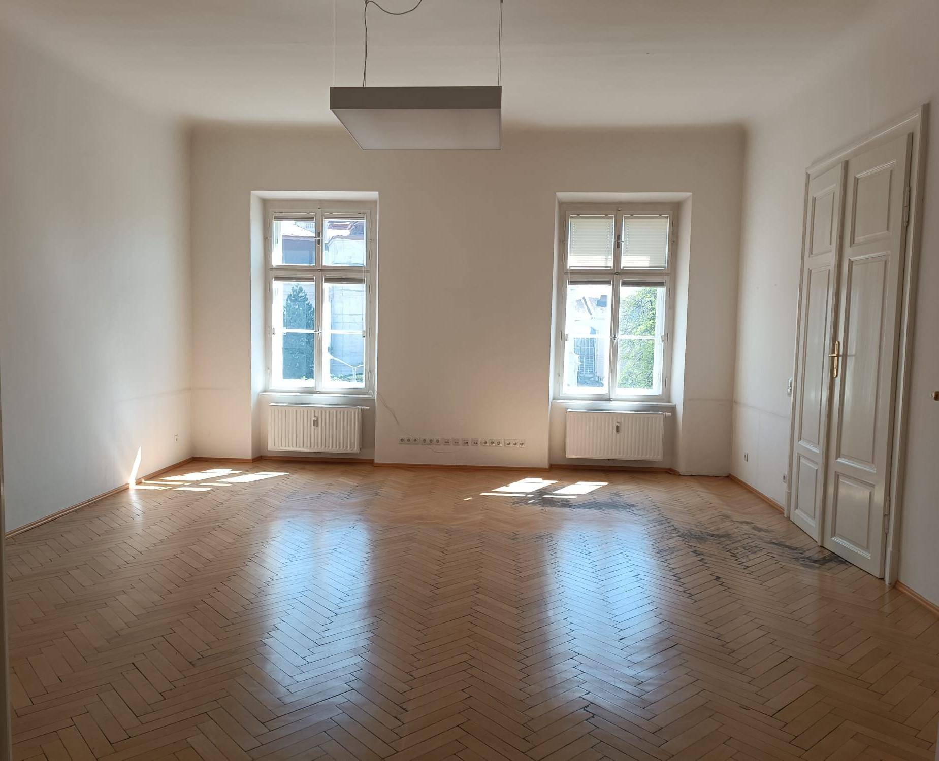 Geräumiger, leerer Raum mit Fischgrätparkett, hohen Fenstern und viel Tageslicht.